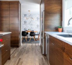 The view from the kitchen into the dining room in the Modern Sensitivity residence. Two large, dark wood-wrapped floor-to-ceiling cabinets line each side of the room. The edge of the stovetop is visible on the right, and the kitchen sink is visible on the left. A brass light fixture hangs in the dining room above a table, and black chairs surround the table. Grey and white cloud-like wallpaper lines the dining room walls.
