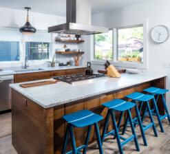 The kitchen in the Modern Sensitivity residence featuring a large, central island wrapped in dark stained wood and finished with a white countertop. Four blue barstools line the island. No upper cabinets.