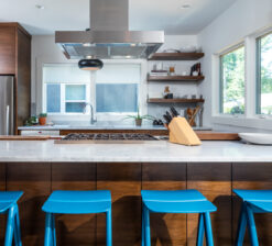 The main island in the Modern Sensitivity kitchen wrapped in dark stained wood and finished with a white countertop. Four bright blue barstools line the island, and a stove vent hood hangs overhead. No upper cabinets.