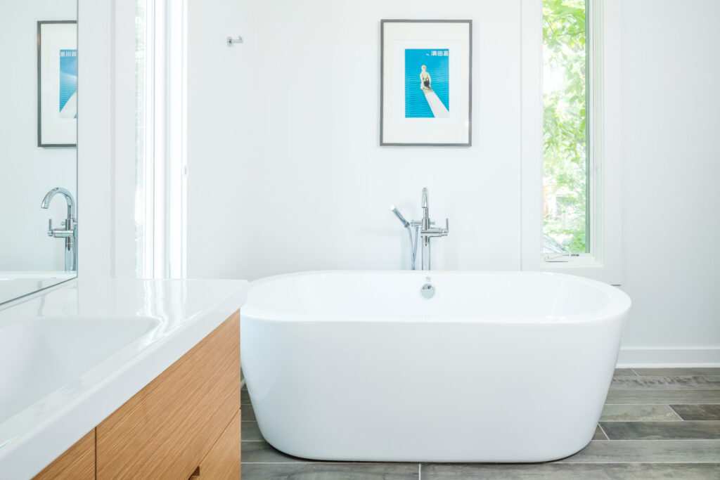 A large, white soaking tub in the primary bathroom of the Modern Sensitivity residence with a single, framed blue print of a woman sitting at the edge of a diving board hung overhead. A skinny vertical window flanks the tub on the right.