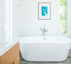 A large, white soaking tub in the primary bathroom of the Modern Sensitivity residence with a single, framed blue print of a woman sitting at the edge of a diving board hung overhead. A skinny vertical window flanks the tub on the right.