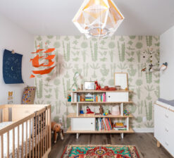 The nursery in the Modern Sensitivity residence with an accent wall covered in a cream and light green wallpaper of flowers and organic shapes. A changing table sits on the left side of the room, and a wood crib to the right. A mid-century book and toy shelf sits in the middle of the room. An orange paper ship hangs from the left corner ceiling.