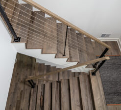 A view looking down from the second level onto the open steel and stringer staircase in the Modern Sensitivity residence with dark grey and brown variegated wood floors and steps.