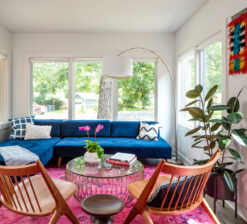A sitting area in the Modern Sensitivity residence lined with large windows on all sides. A navy blue couch sits in the middle of the room with two wood chairs across facing the sofa and a circular glass coffee table in the middle. A large fiddle plant sits near the left wall of the room.