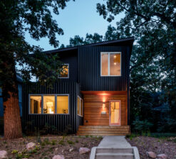 Front exterior view of the Modern Sensitivity home at sunset clad in black siding, accented by warm wood in the entryway. The interior windows are lit.