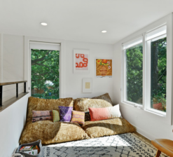 A cozy reading nook on the second level landing of the Modern Sensitivity residence with a large, greenish-yellow reading cushion on the floor. Large windows flank each side of the nook and framed, colorful prints hang on the walls.