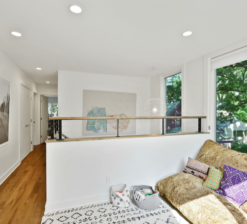 A cozy reading nook connecting to the hallway on the second level landing of the Modern Sensitivity residence with a large, greenish-yellow reading cushion on the floor. The adjacent staircase is open on top. Two large oars are displayed on the left wall next to a framed photograph.