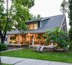 An side exterior view of the Sargent Avenue Redo renovation that now includes a second story addition. Green grass and trees surround the home and the porch and interior lights are lit from the inside.