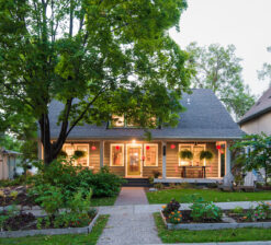 A straight on exterior view of the Sargent Avenue Redo renovation that now includes a second story addition. Green grass and trees surround the home and the porch and interior lights are lit from the inside.
