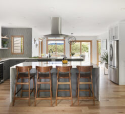 Straight on view of the kitchen in the New Brighton upgrade project. A white island with a waterfall countertop and four dark wood barstools. Grey and white cabinets line the kitchen and an overhead stove vent hood can be seen.