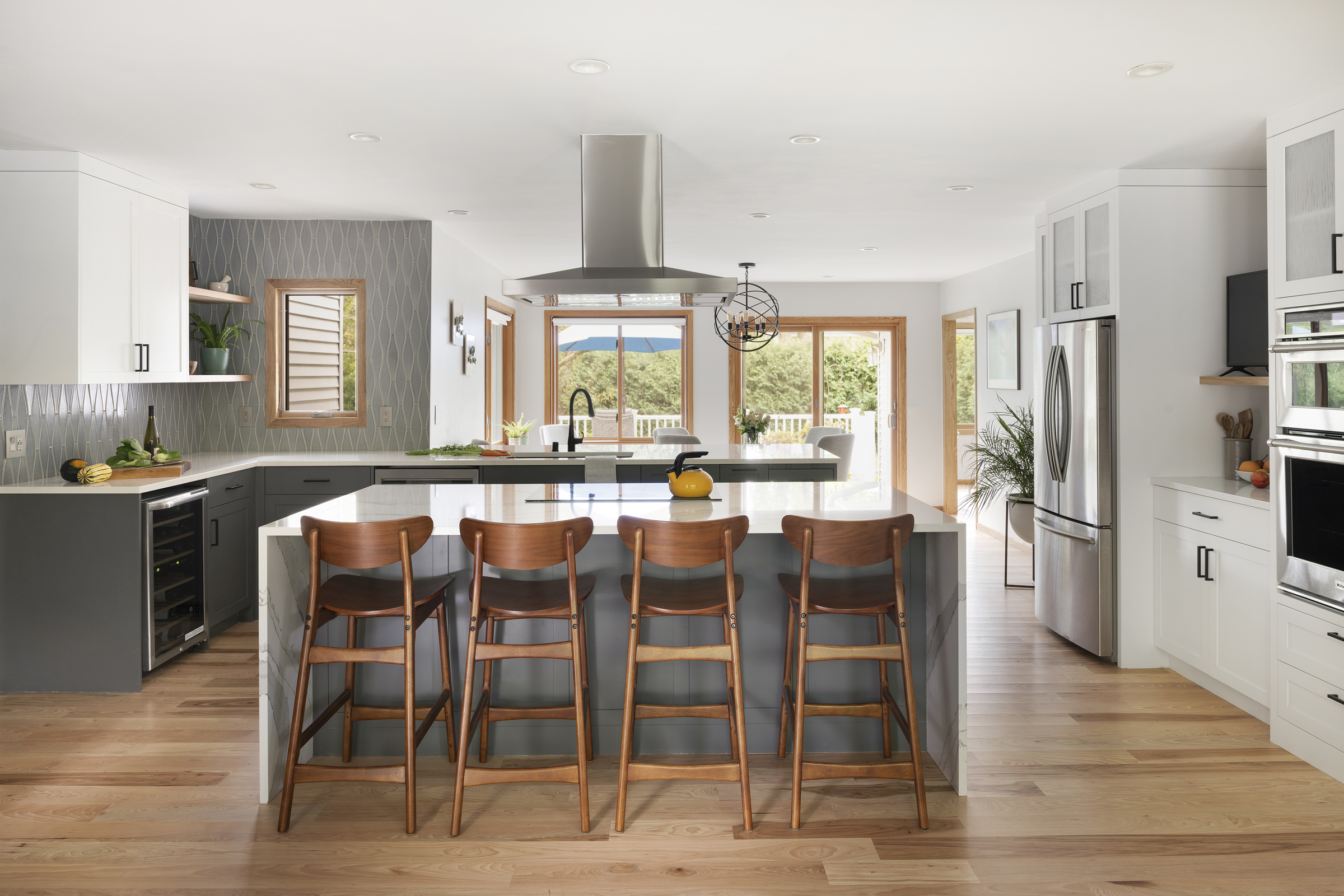 Straight on view of the kitchen in the New Brighton upgrade project. A white island with a waterfall countertop and four dark wood barstools. Grey and white cabinets line the kitchen and an overhead stove vent hood can be seen.
