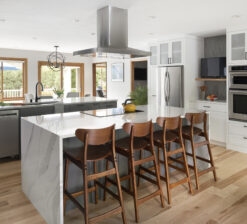 The large central island in the New Brighton Upgrade kitchen renovation with a white waterfall, marble countertop. Surrounding white upper cabinetry and a stovetop vent hood over the island. Four dark wood, high-top stools are pushed into the island.