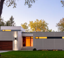 A view of Lakefront Modern's white exterior at golden hour. From this view you can see the wood paneled garage door, front entrance with floor to ceiling glass, and clerestory windows lining the street side perspective of the house.