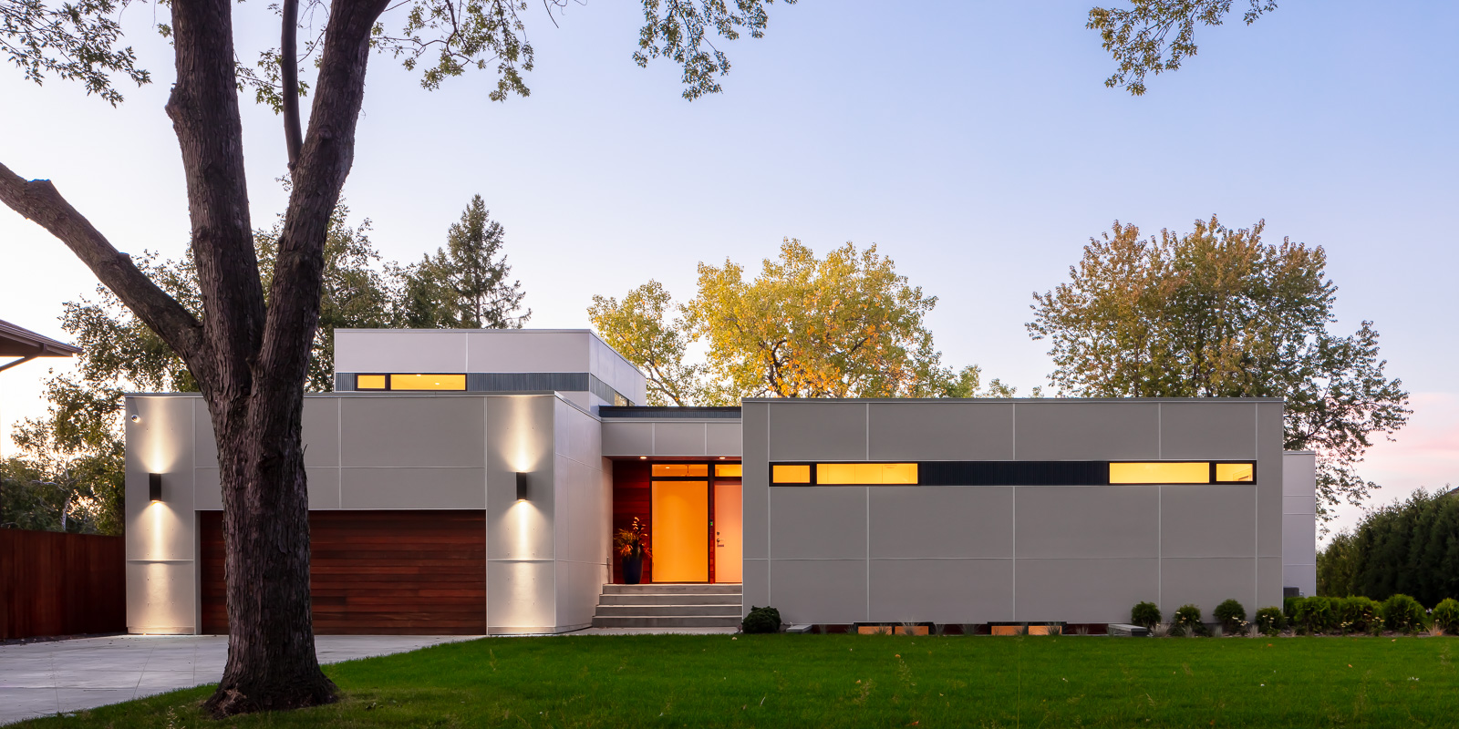 A view of Lakefront Modern's white exterior at golden hour. From this view you can see the wood paneled garage door, front entrance with floor to ceiling glass, and clerestory windows lining the street side perspective of the house.