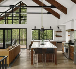 An interior view of the kitchen of the Lakeside Retreat residence. Steel and wood vaulted trusses with a 12 foot sliding pocket door to the lakeside screen porch can be seen beside a large central kitchen island.