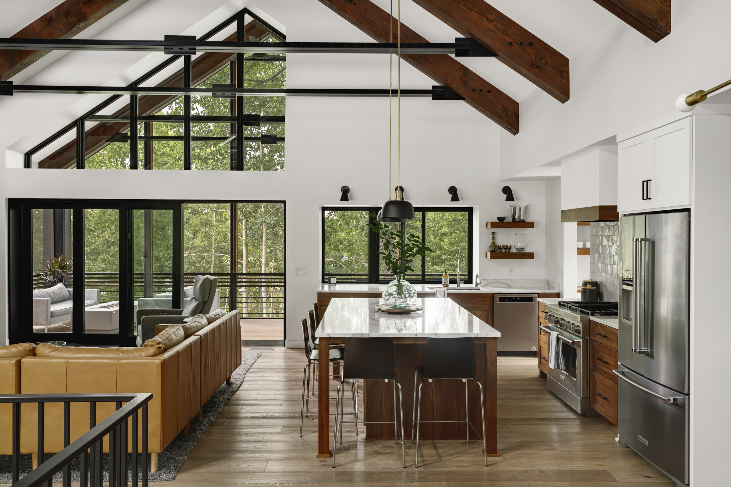 An interior view of the kitchen of the Lakeside Retreat residence. Steel and wood vaulted trusses with a 12 foot sliding pocket door to the lakeside screen porch can be seen beside a large central kitchen island.