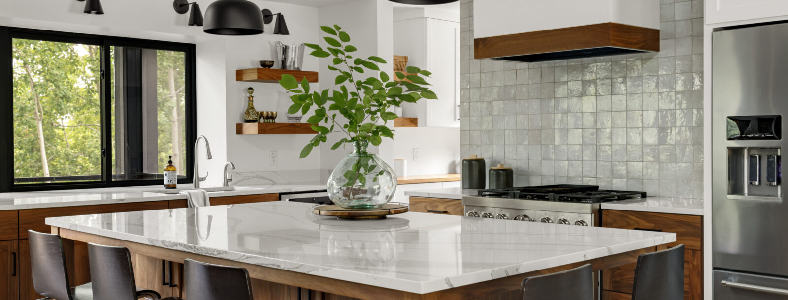 The large kitchen island in the Lakeside Retreat residence with barstools surrounding it on two sides and a large glass vase with cut greens inside sits in the center. An opalescent white tile backsplash lines the wall behind the stove.