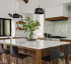 The large kitchen island in the Lakeside Retreat residence with barstools surrounding it on two sides and a large glass vase with cut greens inside sits in the center. An opalescent white tile backsplash lines the wall behind the stove.