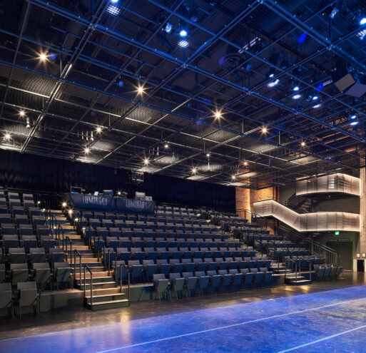 The theater seating, illuminated main staircase, and tension wire lighting grid suspended from the ceiling in the Luminary Arts Center performance space in Minneapolis, MN.