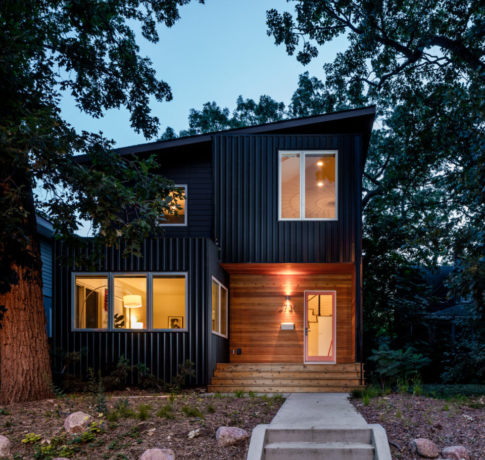 Exterior view of the Modern Sensitivity remodeled residence at dawn with trees surrounding the home on both sides. The interior windows are lit from inside.