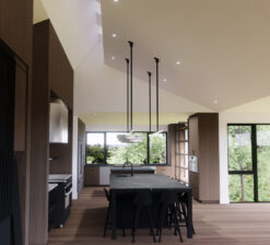 An interior rendering of the kitchen and scullery in the Sirkel Kjøre residence. The large, central kitchen island has a black soapstone countertop, accented by a modern light fixture overhead. Skylight cutouts on the ceiling above let light into the space.