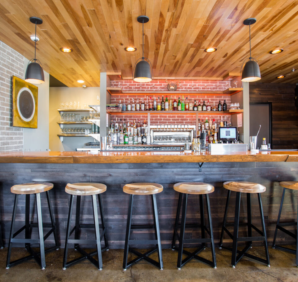The bar at the Oxbow Hotel's The Lakely restaurant with bar stools lined up against it. Wood panels cover the ceiling overhead, black pendant lights hang from above, and a brick red stone backsplash lines the wall behind the bar.