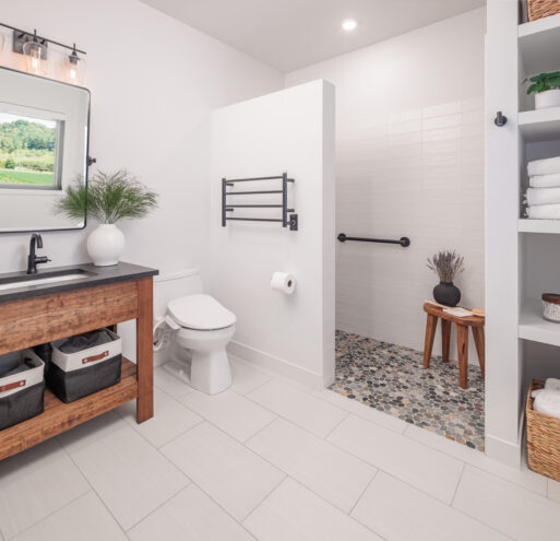 The primary bathroom of the Flower Valley Homestead residence with white walls and matching white tile on the floor, a curb-less shower with a pebble floor, open shelving flanking beside the shower, and a wood vanity.