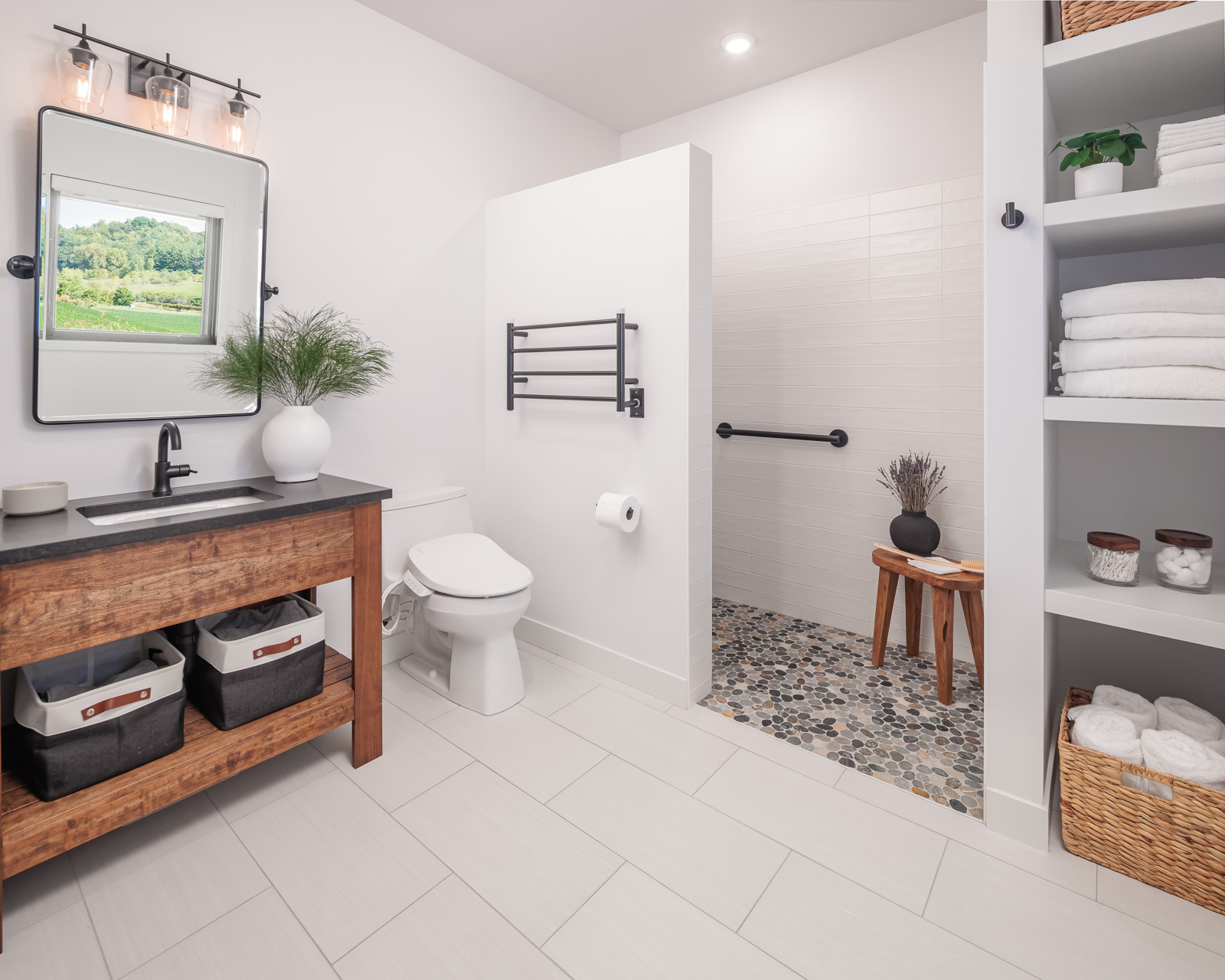 The primary bathroom of the Flower Valley Homestead residence with white walls and matching white tile on the floor, a curb-less shower with a pebble floor, open shelving flanking beside the shower, and a wood vanity.