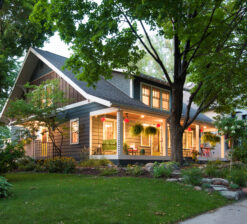 An exterior view of the Sargent Avenue Redo renovation that now includes a second story addition. Green grass and trees surround the home and the porch and interior lights are lit from the inside.