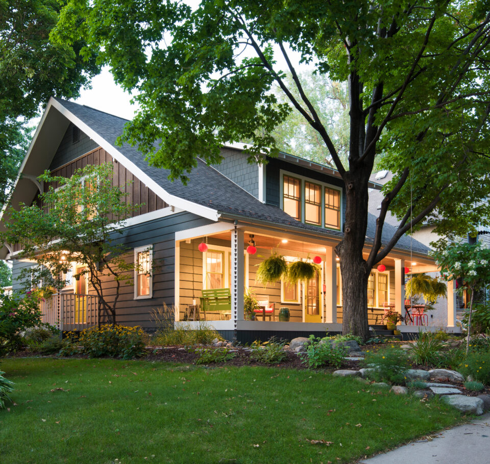 An exterior view of the Sargent Avenue Redo renovation that now includes a second story addition. Green grass and trees surround the home and the porch and interior lights are lit from the inside.
