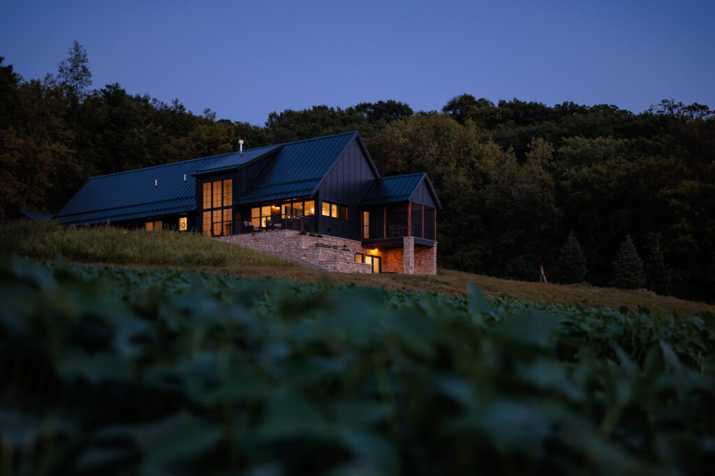 Exterior view of the Flower Valley Homestead residence at dusk in the summer, surrounded by fields of soybeans. The interior windows are lit up. Black siding covers the majority of the home with the bottom lower walkout area covered in limestone.