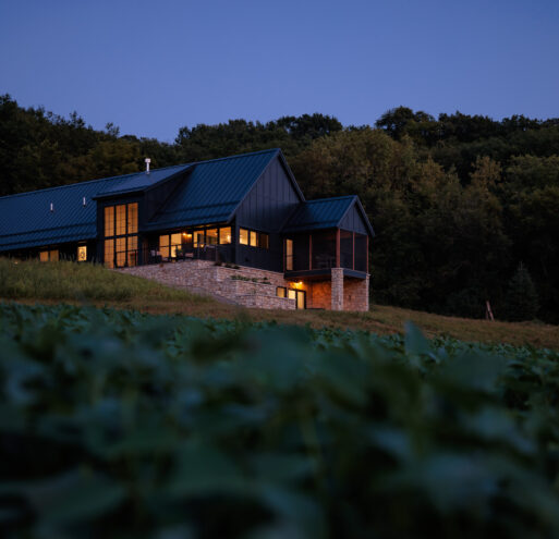 Exterior view of the Flower Valley Homestead residence at dusk in the summer, surrounded by fields of soybeans. The interior windows are lit up. Black siding covers the majority of the home with the bottom lower walkout area covered in limestone.