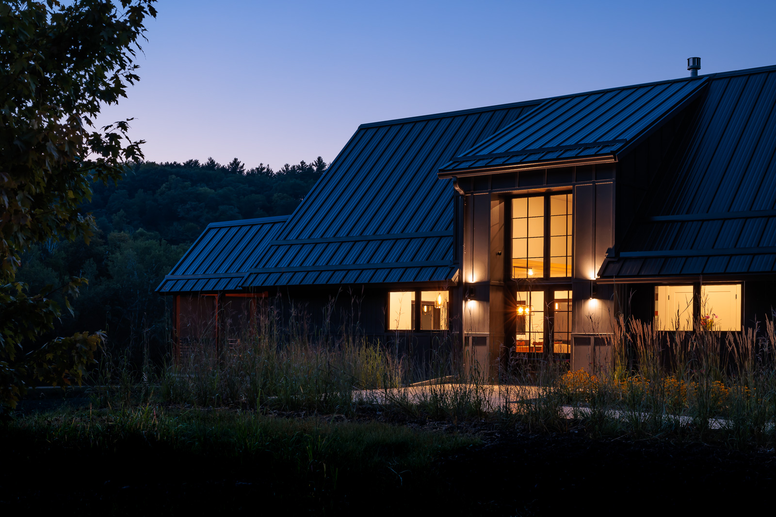A close-up exterior view of the front entrance to Flower Valley Homestead residence just after sun down in the summer. The interior windows are lit up. Black siding covers the entirety of the home in view.