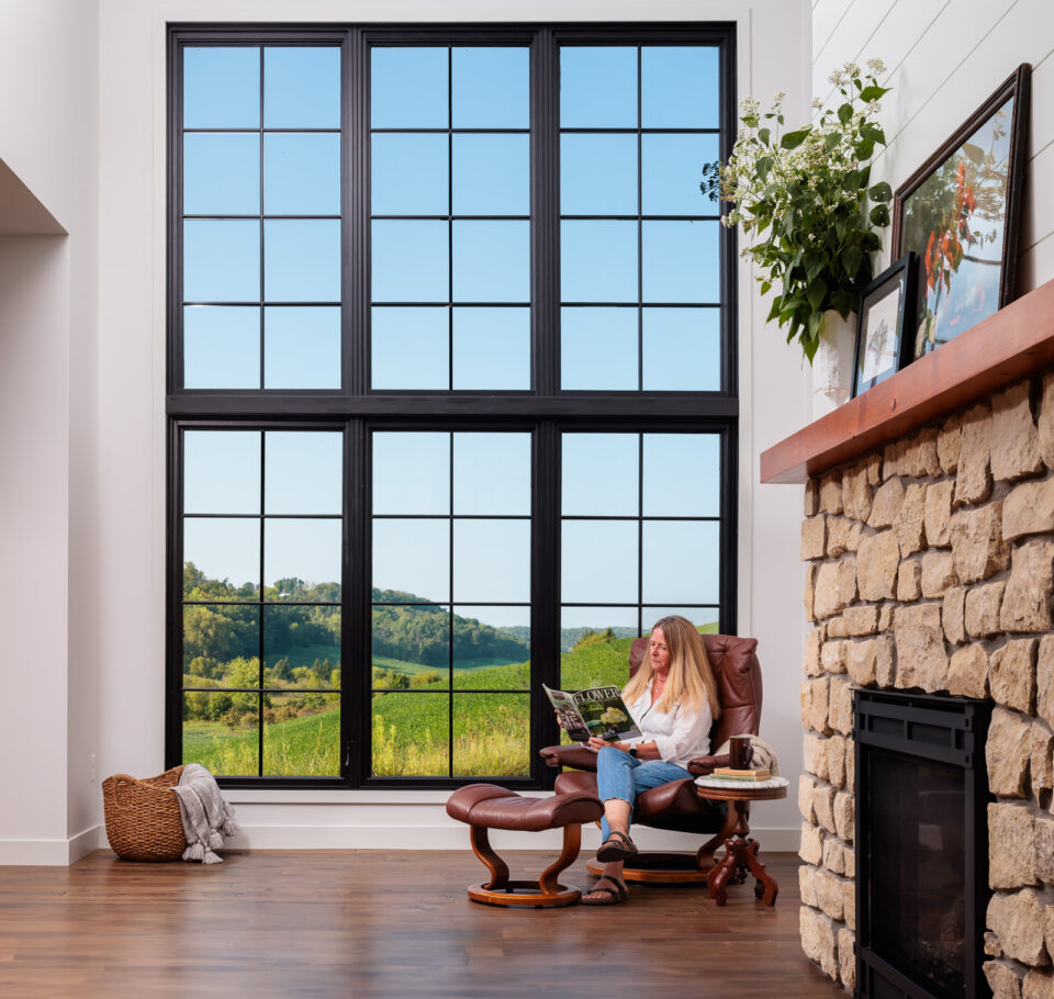 The owner of the Flower Valley Homestead residence reading in her living room chair next to nine foot floor-to-ceiling windows overlooking a valley.