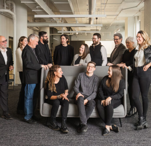 Members of the Shelter Architecture and Interior Design team in their Minneapolis, MN studio. Three people are sitting on a grey couch with more people behind them surrounding it.