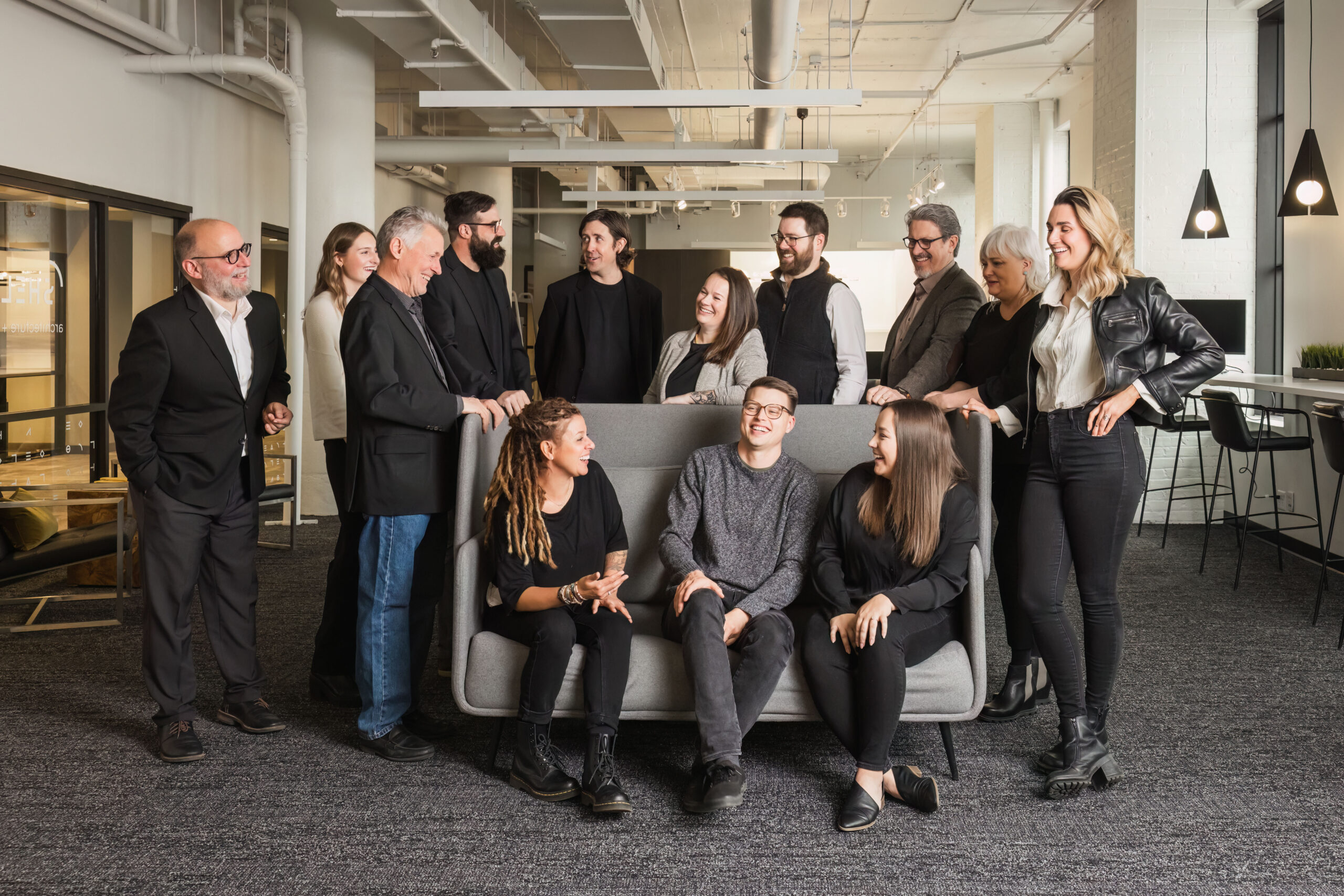 Members of the Shelter Architecture and Interior Design team in their Minneapolis, MN studio. Three people are sitting on a grey couch with more people behind them surrounding it.