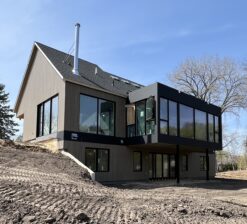 A rear exterior view of the Sirkel Kjøre residence during construction. The multipurpose, glass lined bump-out on the main level of the home overlooks the pond in the backyard. The landscape at this point during construction is bare.