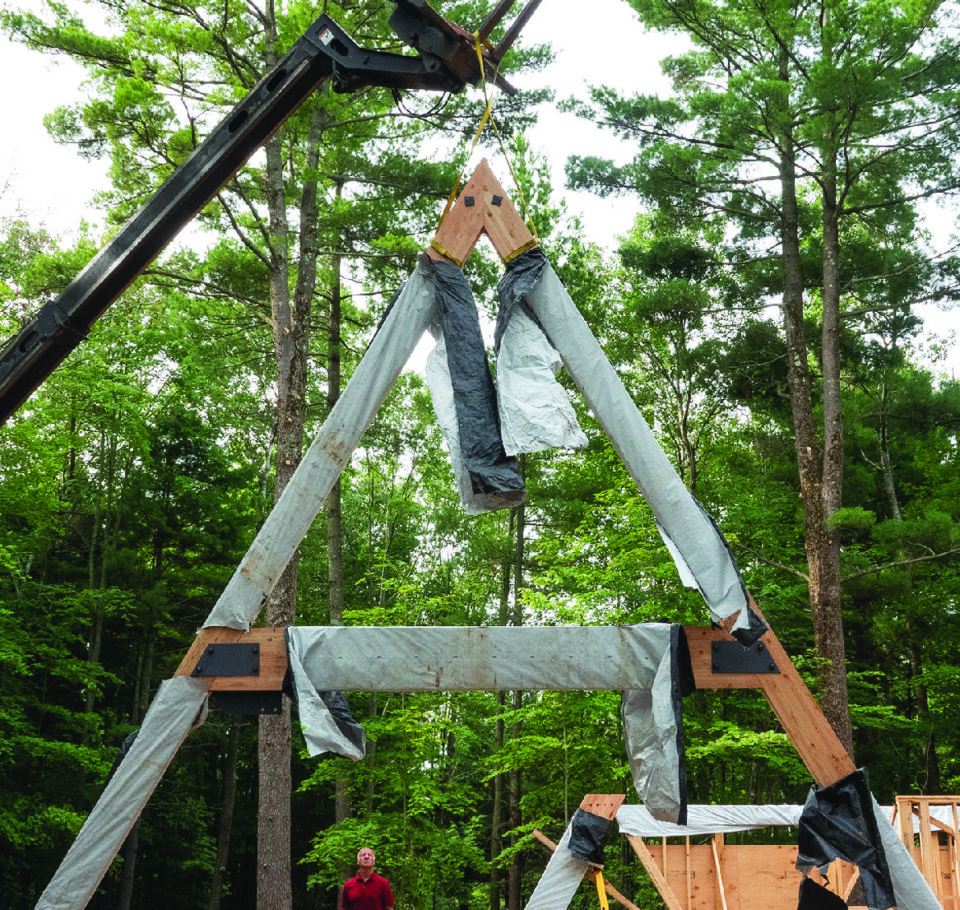 A large triangular wood truss being hoisted into the air during the start of construction on an A-frame house.