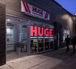 An exterior view of the entrance of the HUGE Improv Theater showing a primary sign spelling out "HUGE" with passerbyers walking past.