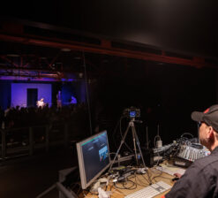 A man seated at the tech booth of the HUGE Improv Theater during a live performance.