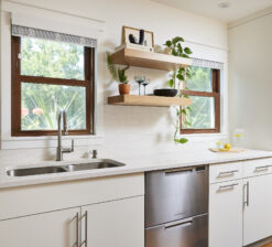 Two open shelves styled with plants and vases sit between two windows in the Saint Paul Bungalow remodeled kitchen. Creamy white lower cabinets are visible and a small built in, drawer dishwasher is as well. A vase with blue and purple flowers sits off to the left.