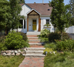 The front exterior of the Saint Paul Bungalow, a one story, bungalow-style home with white siding and a yellow front door. The front entry has a small gabled roof with two beams supporting the overhang. Two small flower boxes sit below two windows on the right side of the home. Landscaping around the home is green and lush.