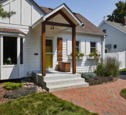 A side exterior view of the Saint Paul Bungalow, a one story, bungalow-style home with white siding and a yellow front door. The front entry has a small gabled roof with two beams supporting the overhang. Two small flower boxes sit below two windows on the right side of the home.